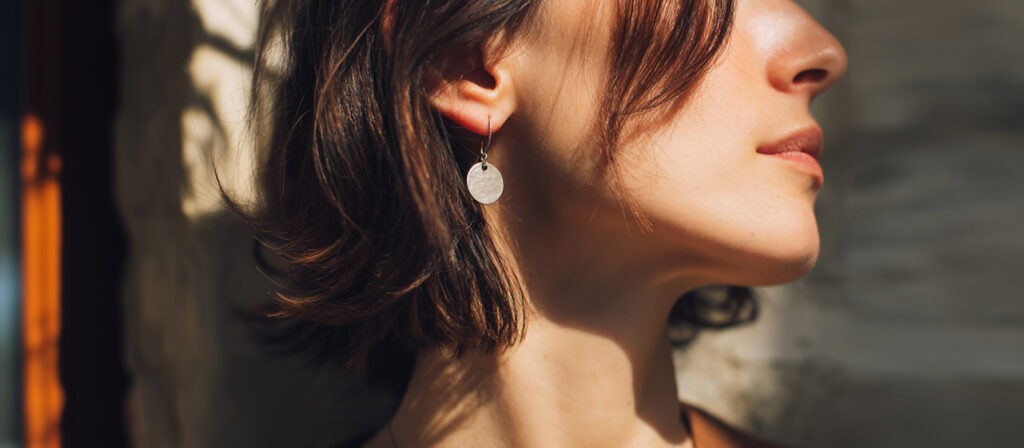 Close-up of a woman wearing a minimalist silver round earring, sunlight highlighting her face and short brown hair.