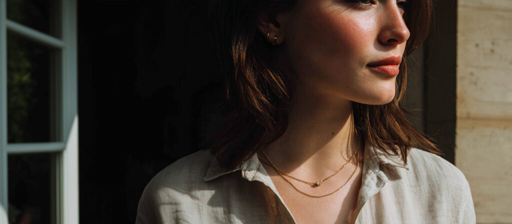 Close-up of a woman wearing delicate layered gold necklaces and small hoop earrings, standing in soft sunlight near a window.