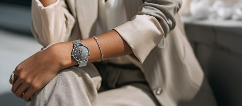 Close-up of a person wearing a silver wristwatch and delicate bracelet, dressed in a beige suit with a relaxed, elegant pose.