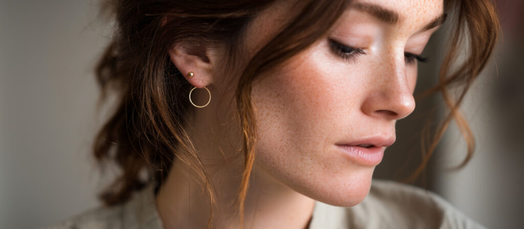 Close-up of a woman with wavy brown hair wearing minimalist gold hoop earrings, softly lit in a neutral indoor setting.